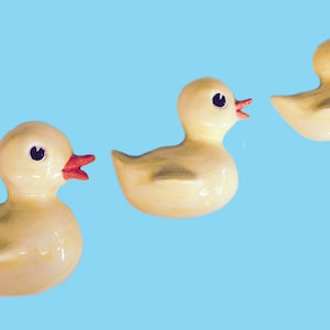 May include: Three yellow rubber duck toys in a row, facing right, against a light blue background. The ducks have black eyes and red beaks.