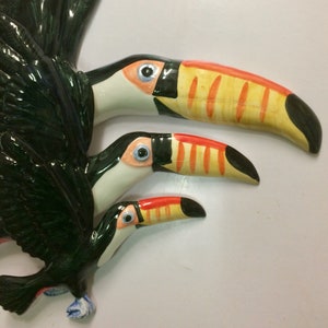 May include: Three ceramic toucans painted in black, white, yellow and orange. The toucans are arranged in a cascading pattern, with the largest toucan at the top and the smallest toucan at the bottom.