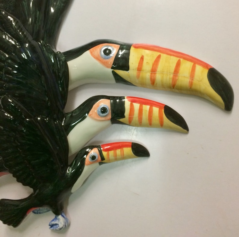 May include: Three ceramic toucans painted in black, white, yellow and orange. The toucans are arranged in a cascading pattern, with the largest toucan at the top and the smallest toucan at the bottom.