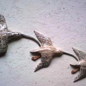 May include: Three wooden sculptures of birds in flight. The birds are painted in shades of brown and white, with intricate details. The sculptures are arranged in a staggered formation, creating a sense of movement.