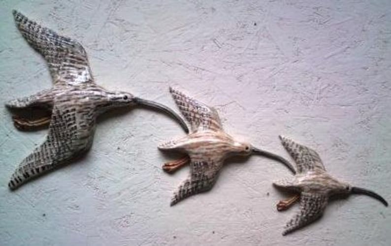 May include: Three wooden sculptures of birds in flight. The birds are painted in shades of brown and white, with intricate details. The sculptures are arranged in a staggered formation, creating a sense of movement.