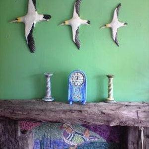 May include: Three white and black bird wall decorations with yellow beaks are mounted on a green wall. Below, a blue clock and two decorative candle holders sit on a wooden mantel. A mosaic design is visible on the fireplace.