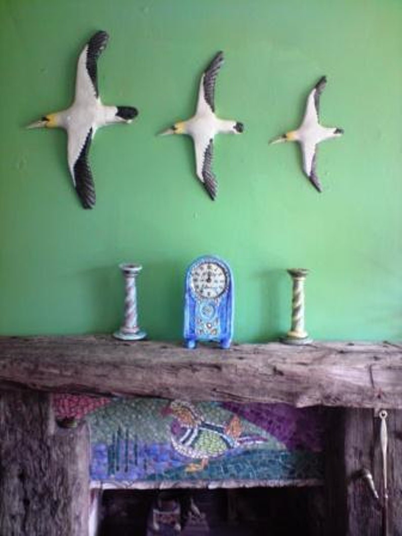 May include: Three white and black bird wall decorations with yellow beaks are mounted on a green wall. Below, a blue clock and two decorative candle holders sit on a wooden mantel. A mosaic design is visible on the fireplace.