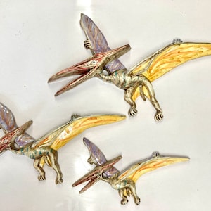 May include: Three ceramic pterodactyl sculptures in various sizes. The largest sculpture is yellow and orange with purple accents. The medium sculpture is yellow and orange with blue accents. The smallest sculpture is yellow and orange with purple accents.