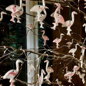 Puede incluir: Una colección de adornos de flamenco de cerámica rosa y blanca colgados de una rama con luces blancas. Los adornos están decorados con un patrón de mármol y purpurina.