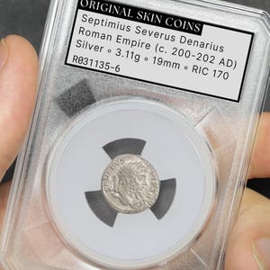 May include: A silver Roman Empire Denarius coin from the Septimius Severus era, c. 200-202 AD, encased in a clear protective holder. The coin is 19mm in diameter and weighs 3.11g. The holder includes a label with details: "ORIGINAL SKIN COINS".