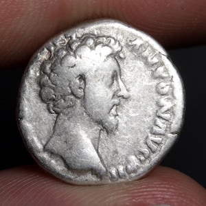 May include: A circular silver ancient coin featuring a profile portrait of a bearded figure facing right. The surface shows signs of wear and age with raised relief details of the head and hair. The edge of the coin displays faint, worn lettering.