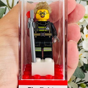 May include: A Lego minifigure firefighter with light brown hair and glasses, wearing a black uniform with yellow reflective stripes, holding a grey tool with a red tip, enclosed in a clear plastic display case with a white base labelled Firefighter Cross.