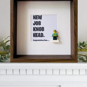 May include: A framed print with a Lego minifigure wearing a yellow vest and a green shirt. The print says "New Job Knob Head. Congratulations Cam..."