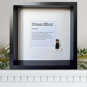 May include: A black shadow box frame containing a white card with the printed definition of Prison Officer [noun] and a small plastic minifigure wearing a dark uniform and cap, standing on a white block labelled with the word Dad in black text.