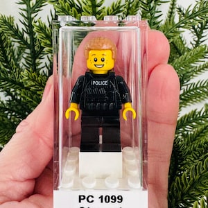 May include: A LEGO minifigure police officer with light brown hair, freckles, and a yellow face, wearing a black tactical vest with the word POLICE printed on the front. The figure stands on a white base inside a clear plastic display case labelled PC 1099 Stewart.