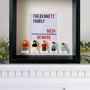 May include: A black framed picture with a white background. The text "THE BENNETT FAMILY NG24 NEWARK" is printed in black and red. Five Lego minifigures are arranged below the text, each with a name tag: Dad, Mum, Leo, Joey, and Kit.