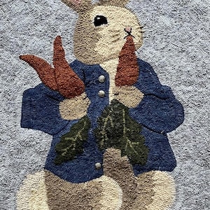 May include: Hand-hooked wall hanging featuring a rabbit in a blue jacket, holding carrots and leaves. The rabbit has tan and white fur, pink ears, and black eyes. The background is a textured gray. Text at the top reads "Hooked By Ruth Poole."