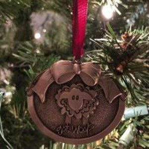 May include: Bronze-colored Christmas ornament with a sheep design and the text "got wool?" A red ribbon is attached for hanging. The ornament is round with a bow at the top and is hanging on a Christmas tree.