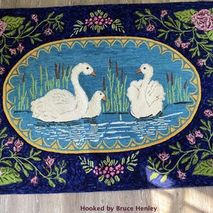 May include: A rectangular hooked rug featuring a central oval panel with three white swans swimming among green cattails on blue water. The dark blue border is decorated with pink roses and green leaves. The text Hooked by Bruce Henley is visible.