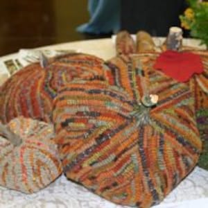 May include: A collection of handmade fabric pumpkins in various sizes and colors, including orange, brown, and green. Each pumpkin features a unique textured pattern and is topped with a small stem and leaf. The pumpkins are arranged on a white surface.