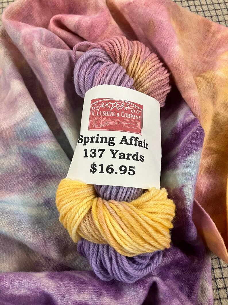 May include: A ball of yarn in yellow, purple, and pink, labelled "Spring Affair 137 Yards" by W. Cushing & Company. The yarn is set against a backdrop of tie-dye fabric in similar pastel colours.