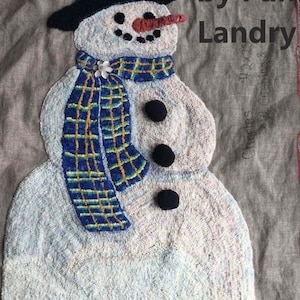 May include: Hand-hooked snowman with a black top hat, carrot nose, and a blue and yellow plaid scarf. The snowman has three black buttons and the text "hooked by Pam Landry".