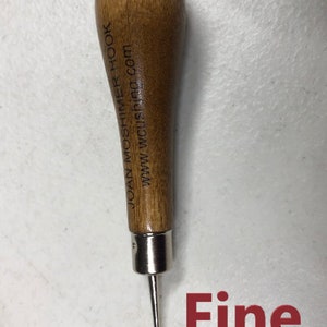 May include: A wooden crochet hook with a metal hook and a brown wooden handle. The handle is engraved with "Joan Moshimer Hook" and "www.wcshor.com". The hook is labeled "Fine".