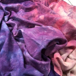May include: A close-up of a soft, purple and pink tie-dye fabric. The fabric is folded and has a gradient of colour from pink to purple.