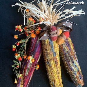 May include: Three decorative corn husks in shades of red, yellow, and purple, adorned with raffia, a brown polka-dot ribbon, and felt acorns. A sprig of orange berries adds a touch of autumn. The text "hooked by Lynn Ashworth" is visible.