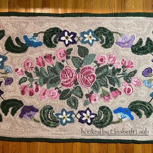 May include: A rectangular hooked rug with a beige background and a dark green border. The design features a central cluster of pink roses and green leaves, surrounded by smaller blue, purple, and pink flowers. The text "hooked by Elizabeth Laub" is visible.