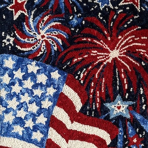 May include: A close-up of a red, white, and blue hooked rug with a patriotic theme. The rug features a design of fireworks and an American flag.