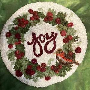 May include: Round, off-white, hand-tufted wall hanging with a wreath design. The wreath features green leaves, red berries, and the word "joy" in burgundy script. A small bird with orange and brown feathers is perched on the wreath.