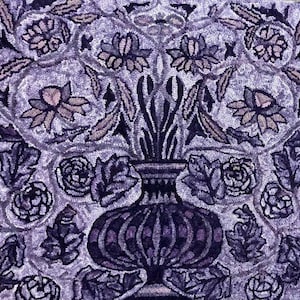 May include: A close-up of a hand-tufted rug with a floral design. The rug features a vase with stylized leaves and flowers in shades of purple, lavender, and gray. The design is symmetrical and detailed, creating a textured appearance.