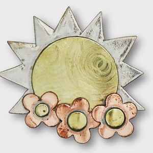 May include: A metal sunburst brooch with a yellow centre and silver rays. Three pink and copper-coloured flowers are at the bottom, each with a yellow centre. The brooch has a rustic, handcrafted appearance.