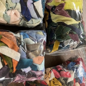 May include: Four clear plastic bags filled with colorful fabric scraps. The scraps are various colors, including blue, green, yellow, pink, and orange. The bags are arranged on a wooden surface.