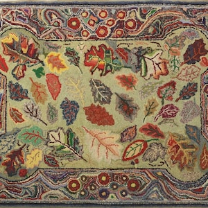 May include: Hand-hooked rug featuring a light green background with a pattern of colorful autumn leaves in shades of red, orange, yellow, and purple. The rug has a decorative border with floral and geometric elements, and a dark blue edge.