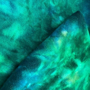 May include: A close-up of a fabric with a tie-dye pattern in shades of teal, turquoise, and green. The fabric is folded over, revealing the texture and color variations. The material appears soft and possibly fleece.