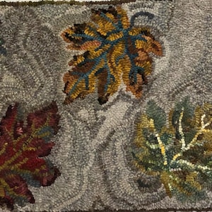 May include: A rectangular rug with a design of autumn leaves in various colours. The leaves, in shades of red, orange, green, and purple, are set against a grey background. The rug is likely made of wool or a similar material.