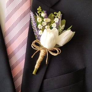 May include: A floral boutonniere featuring two white tulip buds, sprigs of purple lavender, and small white lily of the valley flowers. The stems are wrapped in natural brown twine tied into a small bow, pinned to a black suit jacket with a pink striped tie.