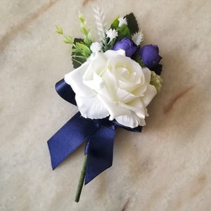 Pode incluir: Uma rosa branca com uma fita azul marinho e pequenas flores roxas. O buquê é perfeito para um casamento ou uma ocasião especial.