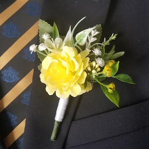 Może przedstawiać: Żółty bukiet do butonierki z białymi i zielonymi akcentami. Bukiet jest przypięty do czarnej marynarki.
