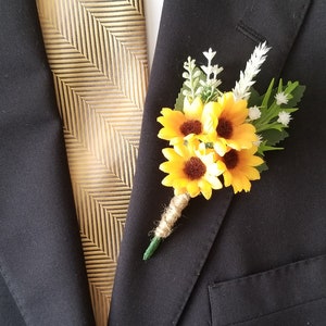 Op de afbeelding: Een gele zonnebloem corsage met groene bladeren en witte bloemen, bevestigd aan een bruine touw strik. De corsage is vastgemaakt aan een zwart colbert.