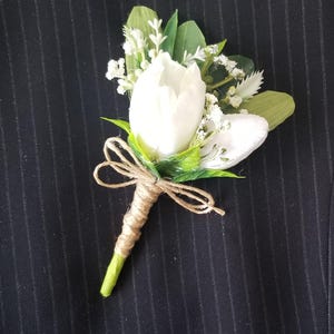 Könnte beinhalten: Ein Ansteckblume mit einer weißen Tulpenknospe, akzentuiert durch kleine weiße Füllblumen und grüne Blätter. Der Stiel ist mit naturfarbenem braunem Garn umwickelt, das zu einer kleinen Schleife gebunden ist, ein dekoratives florales Accessoire.
