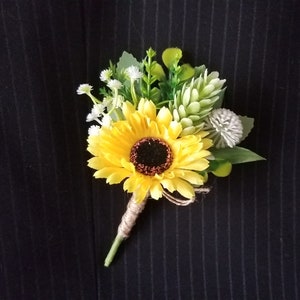 Könnte beinhalten: Ein Ansteckblume mit einer leuchtend gelben Sonnenblume mit dunkelbrauner Mitte, akzentuiert durch kleine weiße Blüten, grünes Blattwerk und ein hellgrünes zapfenartiges Element. Der Stiel ist mit naturfarbenem Bindfaden umwickelt.