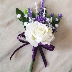Peut inclure: Une rose blanche avec des fleurs violettes et argentées et de la verdure, attachée avec un ruban violet. Cette boutonnière est parfaite pour un mariage ou une occasion spéciale.
