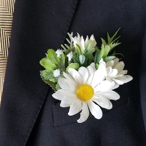 Puede incluir: Una pequeña flor de margarita blanca con centro amarillo y hojas verdes, arreglada en un pequeño ramo, está prendida en una chaqueta de traje negra.