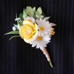 Puede incluir: Un prendido floral con una rosa amarilla y margaritas blancas con centros amarillos, acentuado por hojas verdes y pequeñas flores blancas. El tallo está envuelto en hilo de color beige. El fondo es una tela oscura con rayas finas.