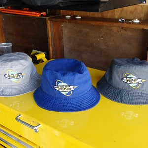 May include: Three bucket hats in different colors: gray, blue, and dark gray. Each hat has a logo with the letters "ISE" in yellow and a surfboard design in blue and yellow.