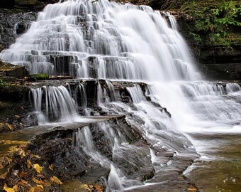 May include: A cascading waterfall flows over layered rock formations. The water is white and foamy, contrasting with the dark, wet rocks and surrounding green foliage. The scene evokes a sense of natural beauty and tranquility.
