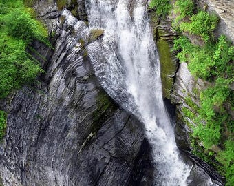 May include: A cascading waterfall plunges down a rugged, layered rock face. Lush green foliage clings to the edges of the cliff, contrasting with the gray and black stone. The water creates a misty spray at the base.
