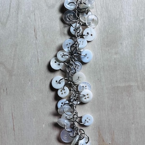 May include: A charm bracelet constructed from a silver-toned metal chain featuring numerous white and translucent buttons of varying sizes attached with individual jump rings. The buttons are circular with two or four holes and a smooth finish.