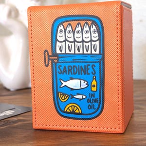 May include: An orange rectangular deck box with a textured surface featuring a graphic illustration of an open tin of sardines. The design includes the text SARDINES IN OLIVE OIL, stylized fish, lemon slices, and a small bottle, all outlined in black.