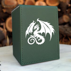 May include: A dark green rectangular deck box featuring a white dragon silhouette printed on the front panel. The box has a textured surface with visible stitching along the edges. A purple twenty-sided die and game cards are visible nearby.