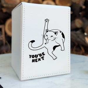 May include: White card storage box with a black and white cat illustration. The cat is licking its paw with the text "YOU'RE NEXT" below. The box is sitting on a white surface with a blurred background of wood.